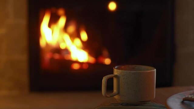 Atmospheric eve at fireplace. Stylish cup of warm tea on wooden table against of cozy burning fireplace. Winter hygge. Autumn cozy evening footage