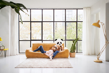 Unrecognizable female couple lying on sofa giant panda bear and sloth stuffed animal masks indoor. Girls in costume posing funny studio photo with natural light and window in background. Copy space.