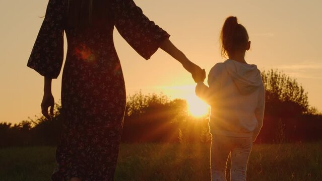 A mother and her daughter walking in a meadow at sunset. silhouettes of a mother and child in the rays of the evening sun. 