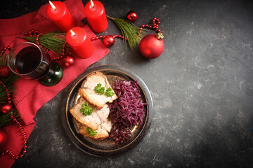 Christmas dinner, roast pork slices with red cabbage on a plate, red candles, wine, napkin and decoration on a dark table, high angle view from above, copy space