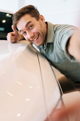 Vertical portrait of cheerful buyer male hugging car hood after purchased in dealership, looking at...