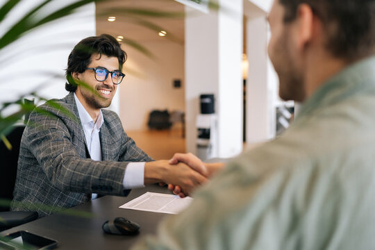 Selective focus of smiling real estate broker in business suit present and advise young man client on decision to sign insurance contract. Happy customer signing purchase agreement and shaking hands.