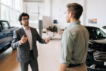 Portrait of professional male dealer in business suit presenting to handsome young man new model of luxury car. Rear view of unrecognizable male customer choosing new vehicle to buy at showroom.