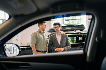 View from interior of car to professional car dealer in business suit selling new car to cheerful male client explaining characteristics. Bearded salesman helping with choice of new auto to guy. - obrazy, fototapety, plakaty