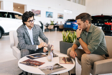 Friendly auto salesman wearing business suit sitting at desk and talking with pensive customer male...