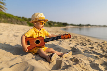 Young baby boy tourist with sunglasses and hat playing ukulele at seaside. Summer vacation concept.