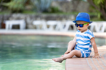 Baby boy wearing sunhat and sunglasses sitting at the edge of swimming pool. Summer relaxation and childhood concept.