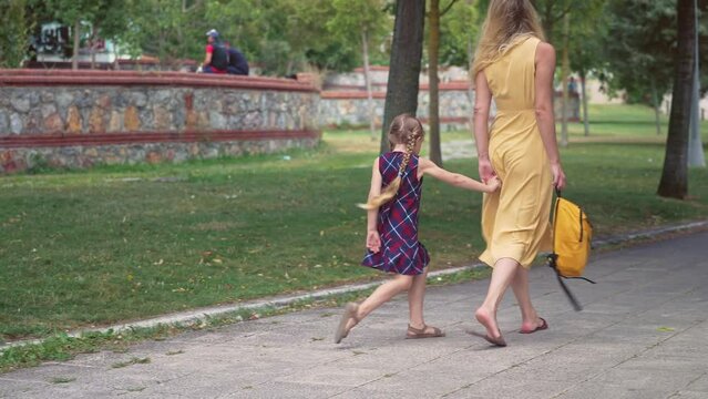 Little girl plays with road tiles walking with mother across green park young woman in yellow summer dress takes active daughter to school in city garden child care