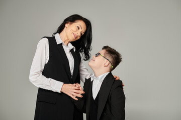 schoolboy with down syndrome and middle aged mother in formal wear smiling at each other on grey