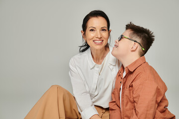 middle aged woman smiling near joyful son with down syndrome on grey, disability acceptance