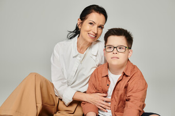 happy and stylish middle aged woman hugging son with down syndrome on grey, supportive parenthood
