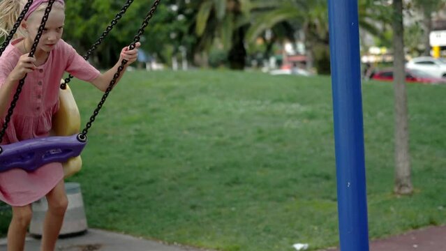 Playful little girl rides safe swings in summer city park happy child in dress enjoys swinging on children playground little kid has fun on attraction