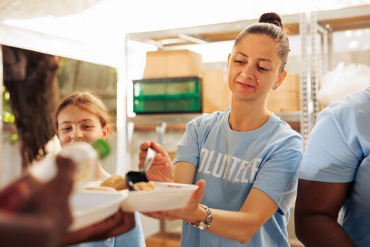Detailed image showcasing female caucasian charity worker serving warm meal to underprivileged african american person. Woman volunteer handing out free food to the hungry and needy homeless people.