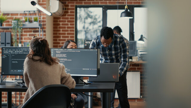 Database programer writing code in front of multiple computer screens displaying artificial intelligence algorithm. Developer coding database while colleagues doing teamwork in background.