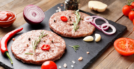 Raw beef hamburger patties on stone plate, vegetables and spices, wooden background.