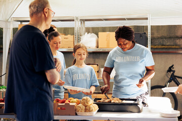 Group of female volunteers at community center distributing free food to people in need. African American woman preparing a meal to give to hungry homeless man at food drive. - obrazy, fototapety, plakaty