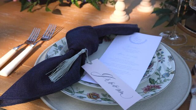 Wedding plate set with floral print, dark-blue napkin and place card for groom. Knife, fork, glasses stand on table decorated for outdoor wedding ceremony. High quality 4k footage