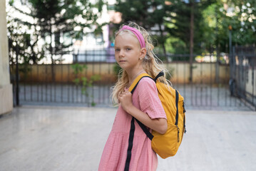 Junior schoolgirl in pink dress stands near school looking at camera girl with long blonde loose hair holds strap of schoolbag child goes to elementary school