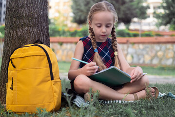 Under a tree, a schoolgirl finds joy in nature while responsibly studying in a green city park on a warm autumn day. 