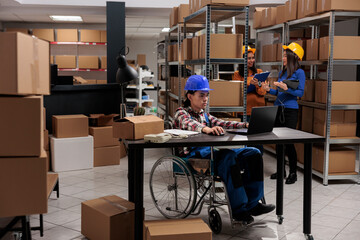 Fototapeta na wymiar Young asian storehouse employee with physical disability managing delivery operations on laptop. Freight distribution center worker in wheelchair doing desk work in warehouse