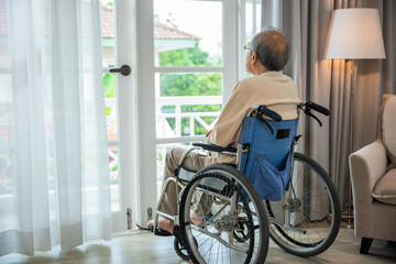 Asian senior man disabled sitting alone in wheelchair looking through window at hospital, lonely elder thoughtful sad old man look outside windows in bedroom at retirement home, feel depressed lonely
