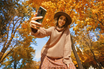Autumn style with this trendy shot. Stylish woman in hat and knit sweater captures the moment with selfie on smartphone at park with orange leaves
