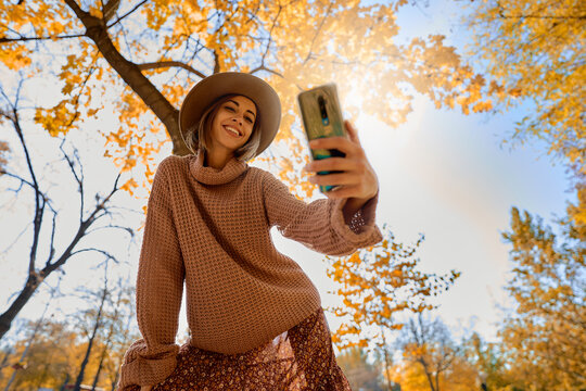 Fall fashion stunning image of woman in hat and cozy knit sweater taking picture on her smartphone at fall park with orange foliage
