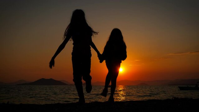 Healthy friends enjoy evening shore. A view of two little sisters silhouettes running and playing on the beach during nightfall. A concept of happy children vacation.
