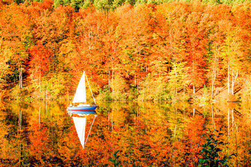 a sailboat and an autumn forest of red yellow and green autumn colors are reflected in the lake surface
