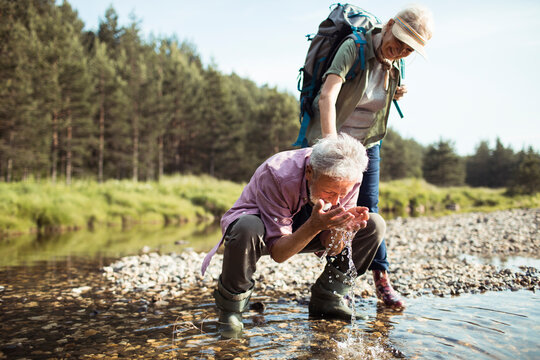 Senior Couple Using The Cold Spring Water To Refresh Themselves While Hiking And Camping In The Forest In Nature