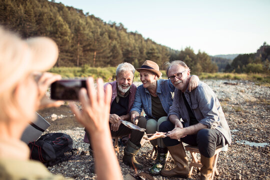 Senior people taking a picture with a smartphone while camping and cooking fish they caught fishing in the creek in the forest