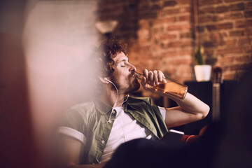 Young man relaxing and drinking a cold beer while listening to music on his smartphone on the couch in the living room
