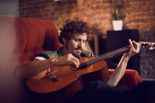 Young caucasian man playing a guitar on the couch in the living room