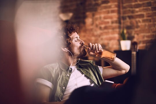 Young man relaxing and drinking a cold beer while listening to music on his smartphone on the couch in the living room