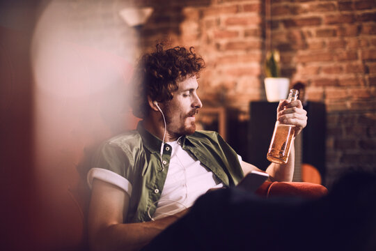 Young man relaxing and drinking a cold beer while listening to music on his smartphone on the couch in the living room