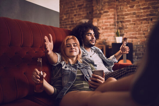 Happy young couple listening to music on the headphones together while having beer on the couch in the living room