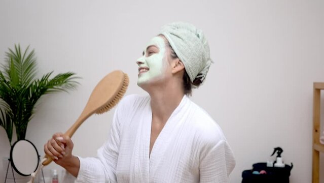 Happy woman with clay mask on face singing in hairbrush mic and dancing at home lady in bathrobe with towel on head having fun on pampering day skincare treatment