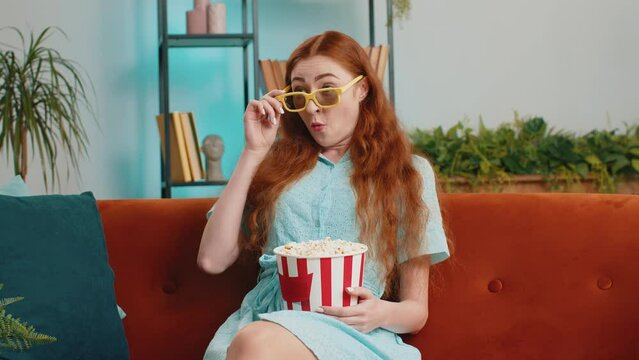 Excited redhead woman sitting on sofa eating popcorn and watching interesting tv serial, sport game, film, online social media movie content at home. Girl in 3D glasses enjoying domestic entertainment