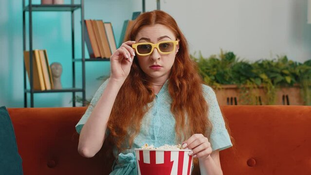 Excited young woman sitting on sofa eating popcorn and watching interesting tv serial, sport game, film, online social media movie content at home. Girl in 3D glasses enjoying domestic entertainment