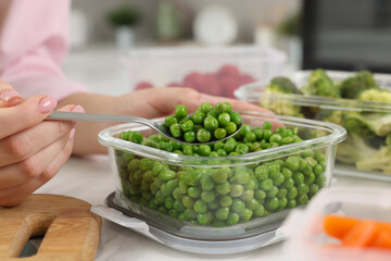Woman taking green peas with spoon from glass container at table in kitchen, closeup. Food storage