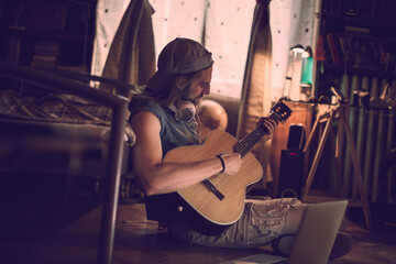 Young man playing the guitar and using a laptop in a bedroom at home