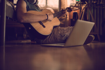 Young man playing the guitar and using a laptop in a bedroom at home