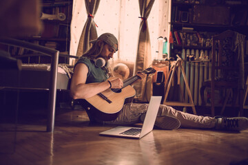 Young man playing the guitar and using a laptop in a bedroom at home