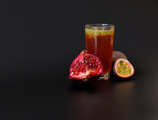 A glass of fruit juice with seeds on a black background, along with a broken pomegranate fruit and pieces of ripe passion fruit.