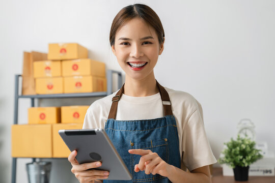 Startup small business, Young Asian woman checking online order on digital tablet and packing boxes for products to send to customers. working at the home office.