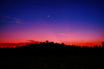 Moon and stars over forest on red and blue of night sky.