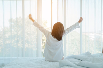 Photo of young happy woman in pajama stretching her arms and smiling while sitting on bed after...