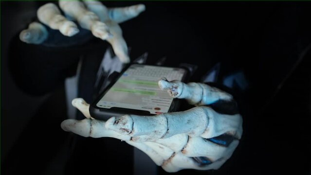 Hands in creepy death costume uses smartphone views and writes in messenger on isolated black background. Halloween party. Online predators.