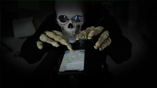 Gloomy man with mask of skull in black poking his bony fingers on smartphone in black background. Fraud in internet. Advertising scary game. Close-up.