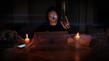 Mysterious man in scary skeleton costume show rocker symbol hand sitting table of laptop with cobwebs and candles in dark room, vignette. Advertising scary game.